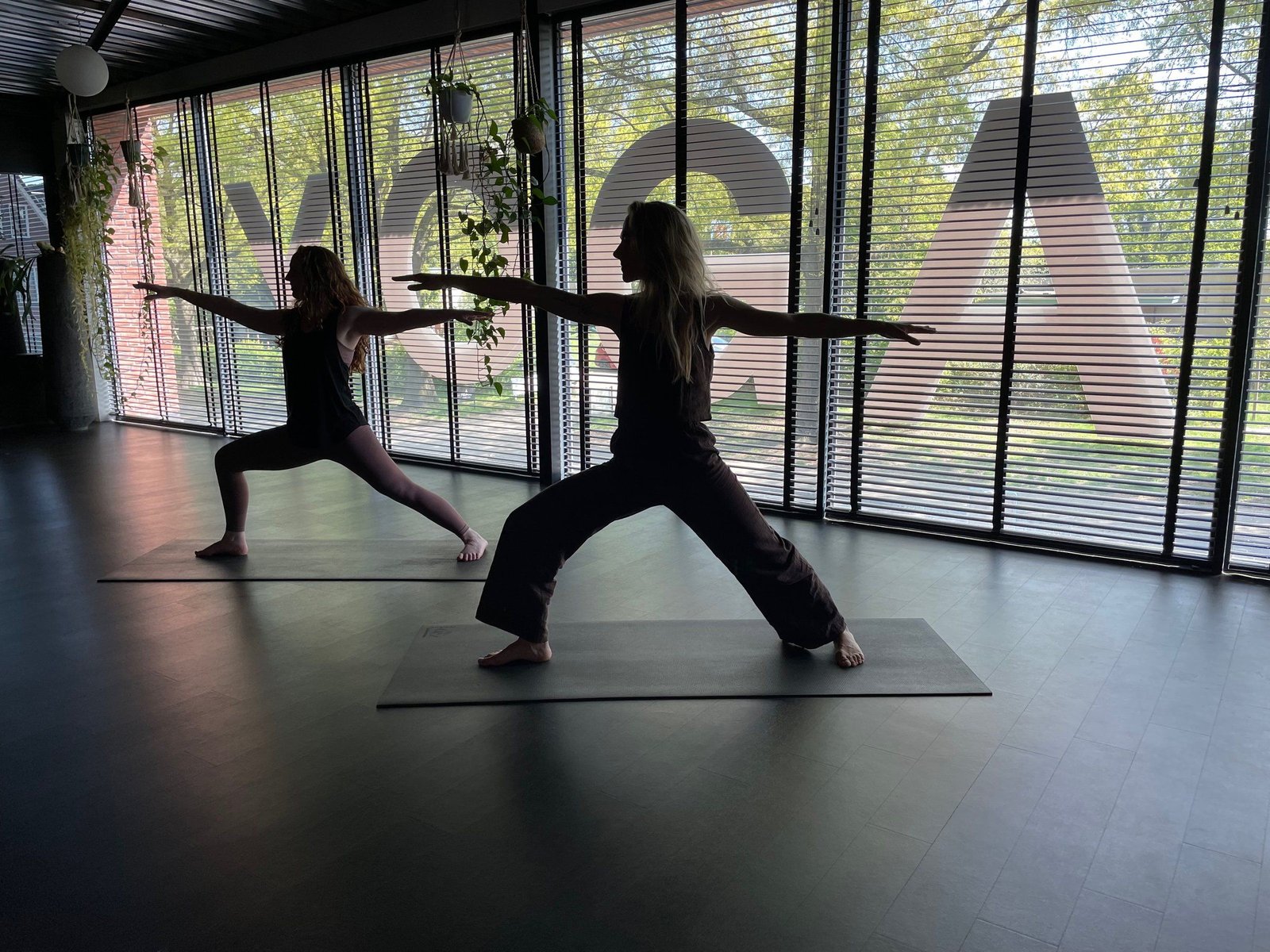 Twee yogi's in de krijgerhouding voor het raam van de grote zaal, met de letters YOGA op het glas